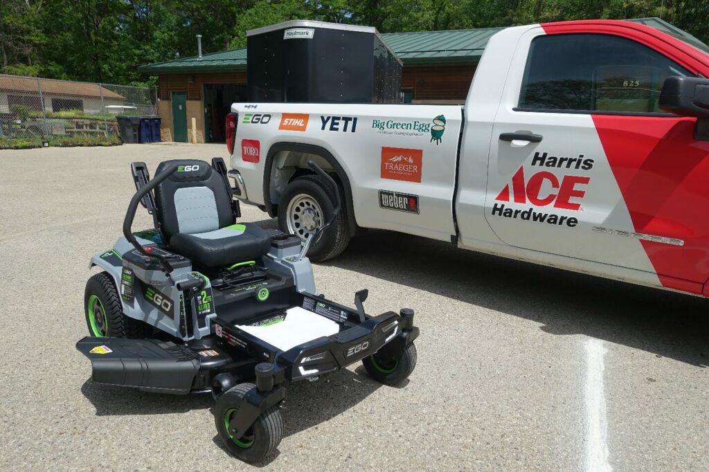 A battery-powered riding lawnmower sits next to a pickup truck from Ace Harware.